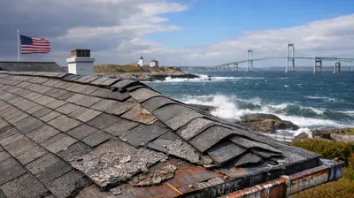 Weather-worn roof of a coastal home in Jamestown, Rhode Island, showing shingle erosion and rusted metal flashing with the Newport Bridge and ocean waves in the background.