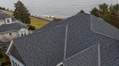 Drone view of a Rhode Island home with a dark architectural shingle roof, multiple valleys, complex rooflines, and a stone chimney.