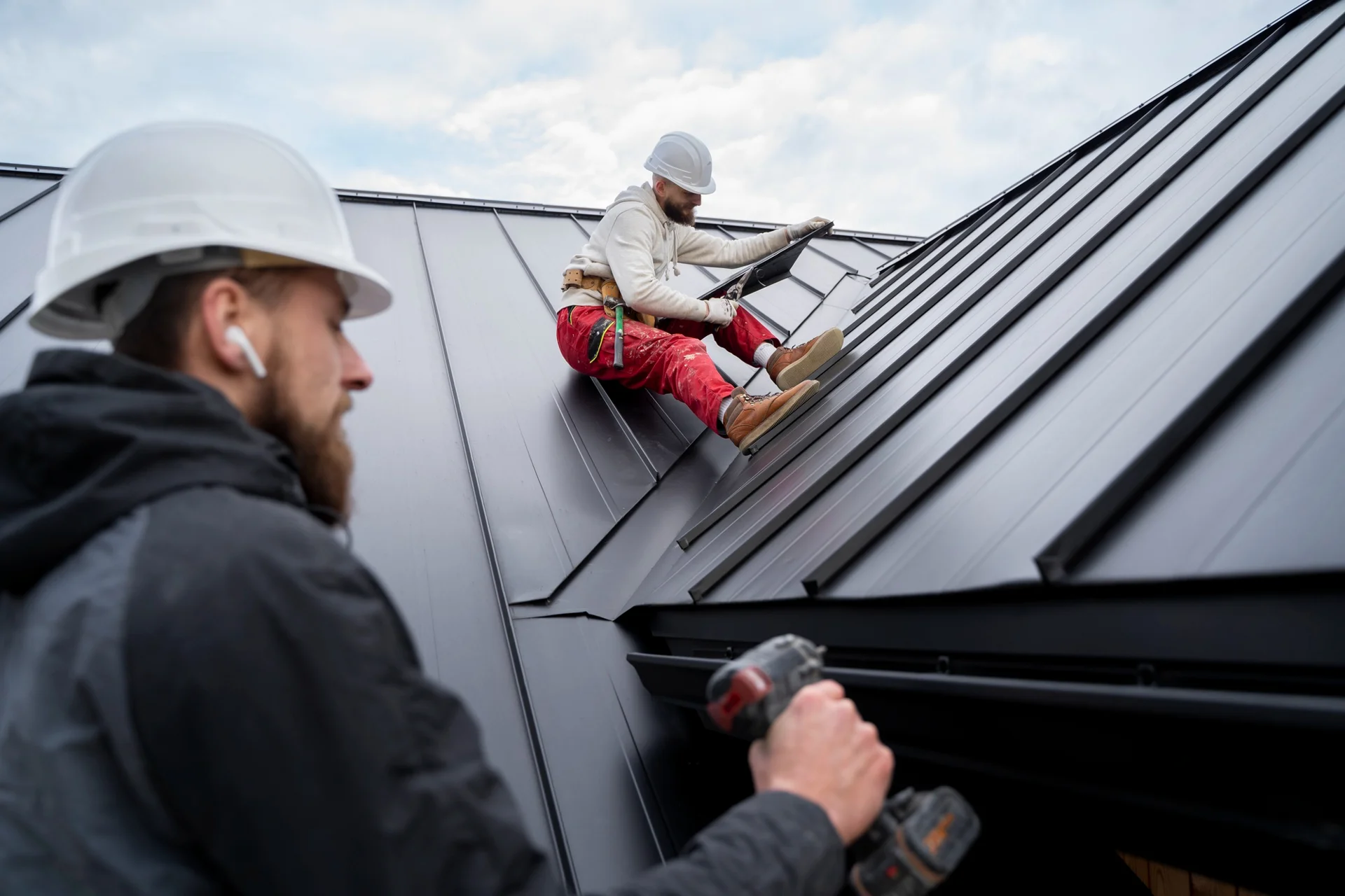 Two construction workers wearing hard hats install a commercial roofing panel in Sarasota, one using a drill while the other positions the panel.